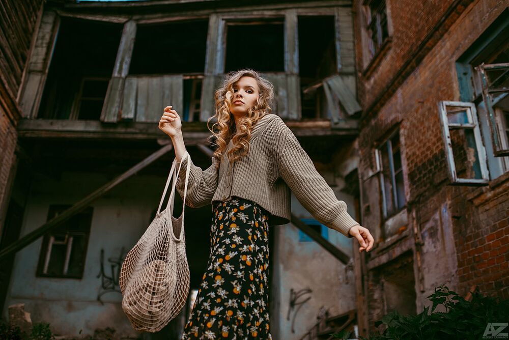 Обои для рабочего стола Модель Полина Белоусова с вьющимися волосами, в кофте, в цветастом платье и с сетчатой сумкой на руке стоит во дворе полуразрушенного дома