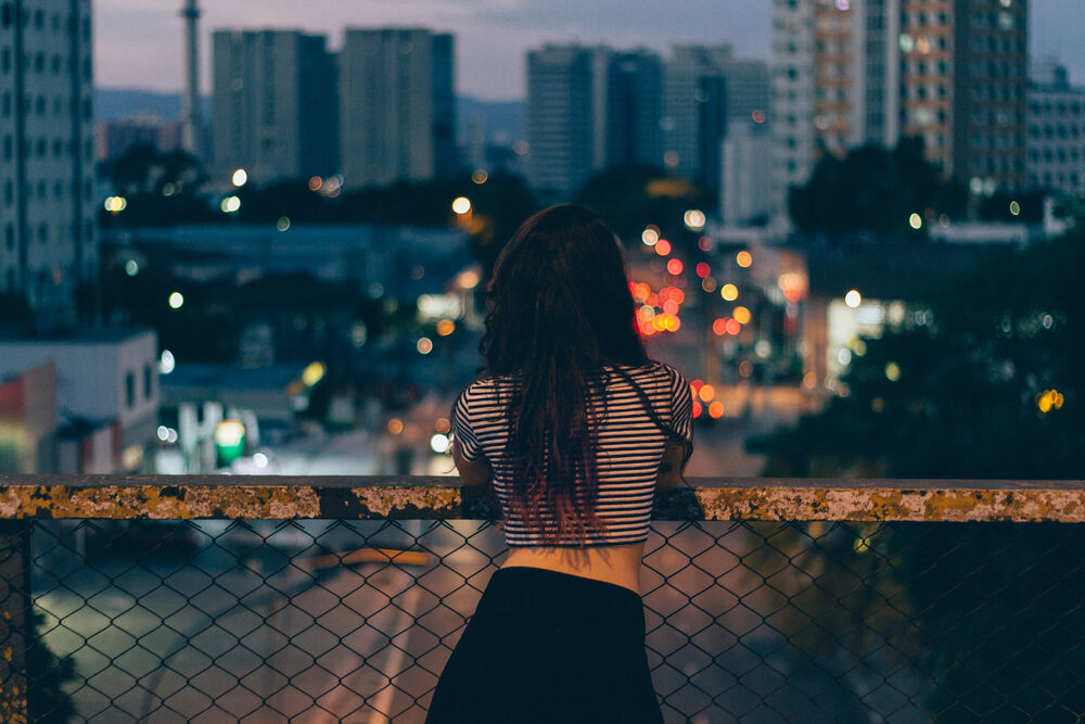 Обои для рабочего стола Девушка в полосатой футболке и черной юбке стоит спиной глядя на вечерний город