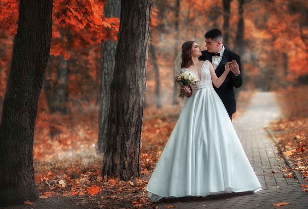 Обои для рабочего стола Мужчина жених в черном смокинге с бабочкой и девушка невеста в белом длинном платье стоят на дорожке в осеннем паке