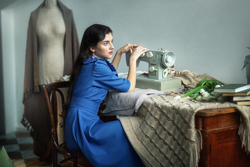 Обои для рабочего стола Брюнетка - модель Злата Зареева в синем платье сидит на стуле в комнате у стола со стоящей на нем швейной машинкой