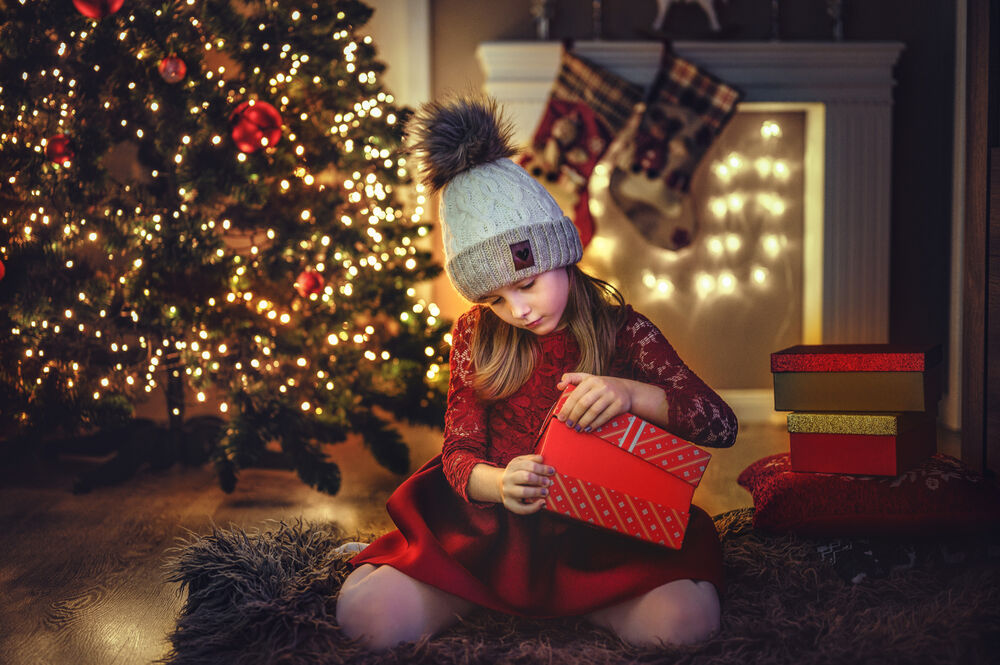 Обои для рабочего стола Девочка в шапочке в комнате с новогодней елкой сидит на полу и открывает подарок
