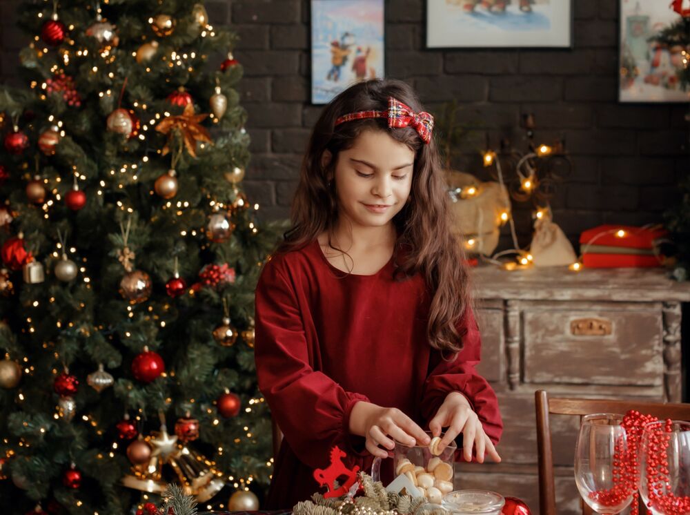Обои для рабочего стола Девочка стоит у стола, держа в руке печенье, в комнате с новогодней елкой