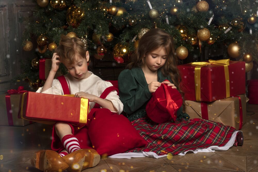 Обои для рабочего стола Мальчик и девочка сидят на полу комнаты у новогодней елки и рассматривают коробки с подарками