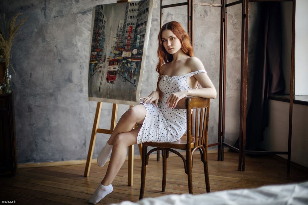 Обои для рабочего стола Модель Надежда Третьякова в белом платье в горошек сидит на стуле в комнате рядом с картиной