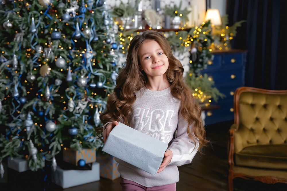Обои для рабочего стола Девочка с подарком в руках стоит на фоне новогодней елки