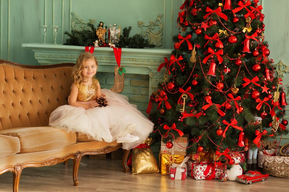 Обои для рабочего стола Девочка в нарядном платье сидит на диване рядом с новогодней красивой елкой и подарками под ней