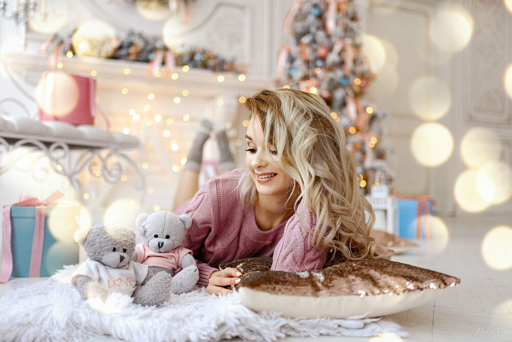 Обои для рабочего стола Белокурая девушка в розовом свитере лежит на постели с игрушечными мишками в комнате с новогодней елкой