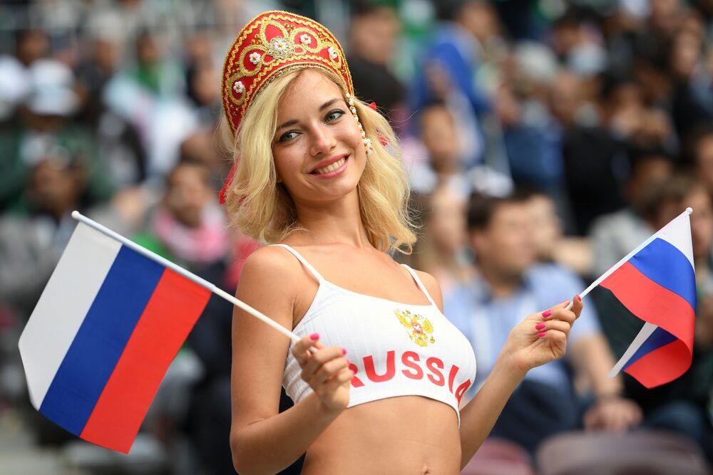 Обои для рабочего стола Девушка Наталья Немчинова - Андреева в кокошнике с красной надписью Russia на груди и двумя российскими флагами в руках на фоне людей