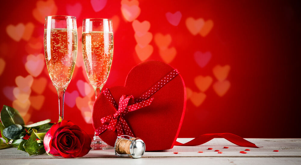 Обои для рабочего стола Два бокала с шампанским, красная роза, пробка и подарочная коробка в виде сердечка, перевязанная лентой на красном фоне с сердечками
