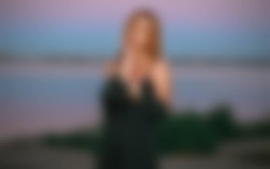 Обои на рабочий стол Рыжеволосая девушка в приспущенном черном платье стоит на размытом фоне природы и водоема