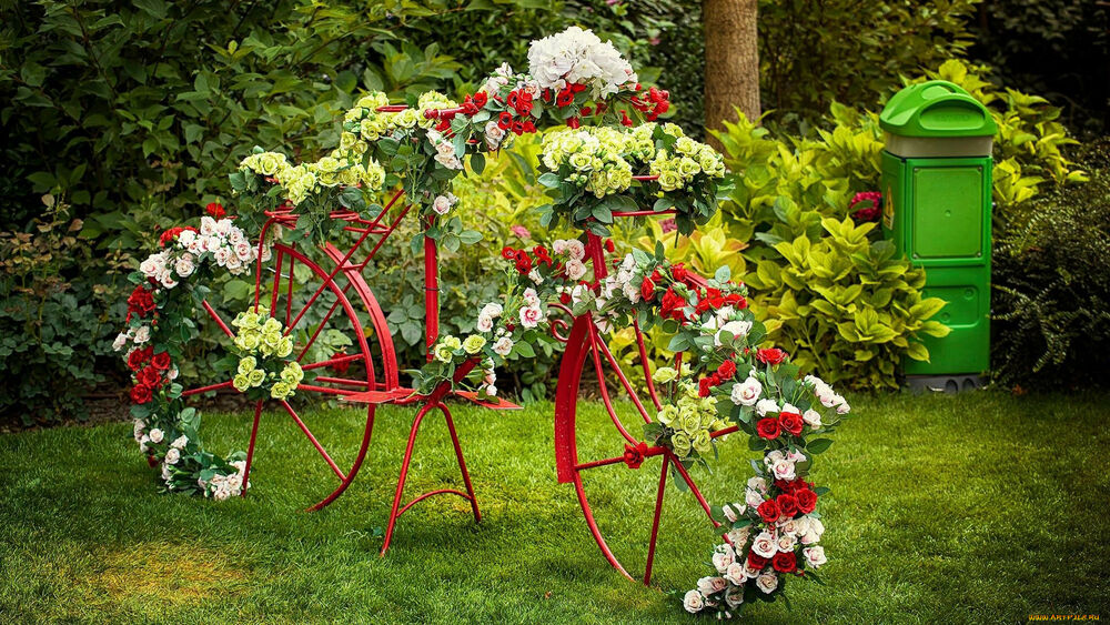 Обои для рабочего стола Старинный велосипед красного цвета стоит на лужайке украшенный букетами искусственных цветов, на фоне кустарников и деревьев