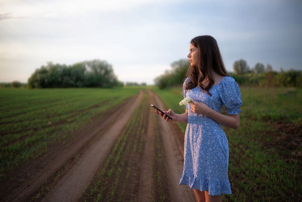 Обои для рабочего стола Модель Валерия в легком голубом платье со смартфоном в руке стоит на дороге в поле