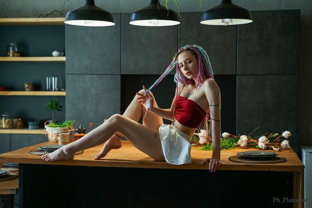 Обои для рабочего стола Модель Vika с разноцветными косичками, в бордовом топе и в белой юбке сидит на столе на кухне