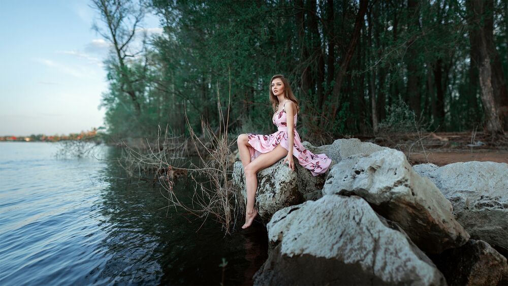 Обои для рабочего стола Модель Veronica в розовом платье сидит на камнях на берегу водоема
