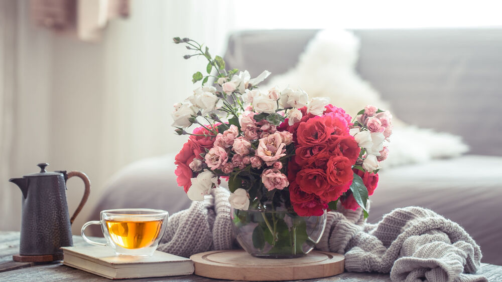 Обои для рабочего стола Букет цветов стоит на столе с чашкой чая книгой и чайником на фоне серого дивана