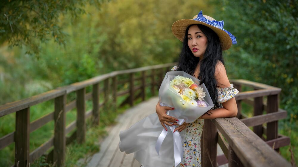 Обои для рабочего стола Модель Ева в шляпе и в платье в цветочек стоит на мосту на фоне природы с букетом цветов в руках