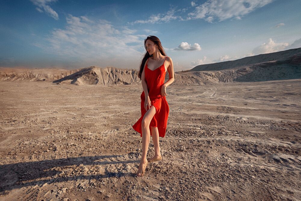 Обои для рабочего стола Брюнетка в красном длинном платье стоит на пустынной холмистой местности на фоне неба и облаков