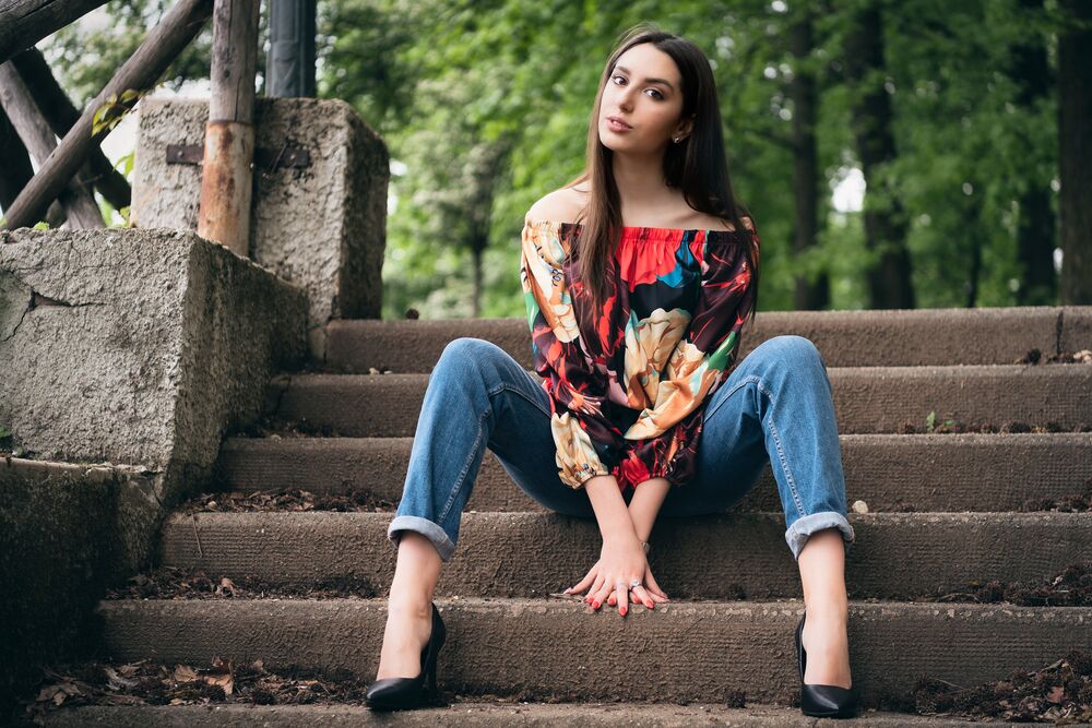 Обои для рабочего стола Модель Alessandra в цветастой блузке и джинсах сидит на ступеньках лестницы на фоне природы