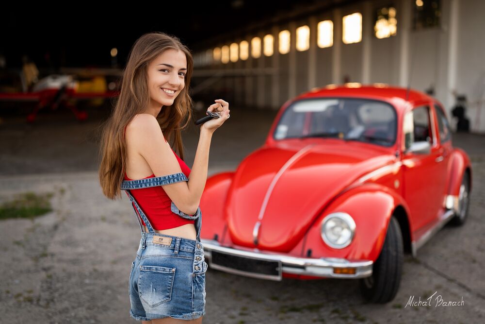 Обои для рабочего стола Модель Weronika в красном топе и в джинсовых шорах стоит в ангаре рядом с авто Volkswagen Beetle