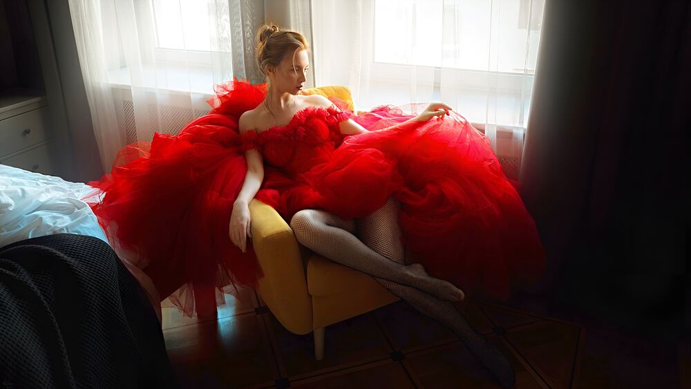 Обои для рабочего стола Блондинка в красном платье с декольте сидит в кресле у окна в спальне