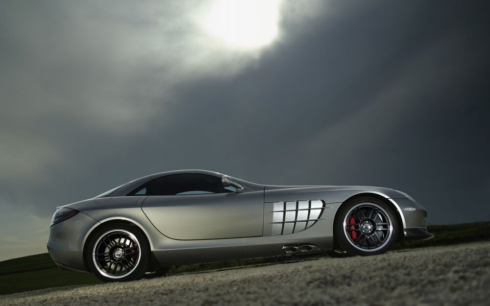Обои для рабочего стола Серебристый спортивный автомобиль Mercedes-Benz SLR McLaren едет по холмам на фоне пасмурного неба