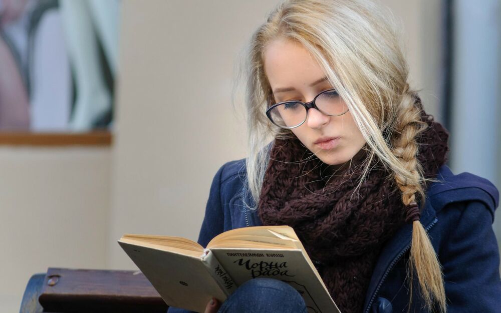 Обои для рабочего стола Блондинка в темно-синей одежде с коричневым шарфом в очках читает книгу