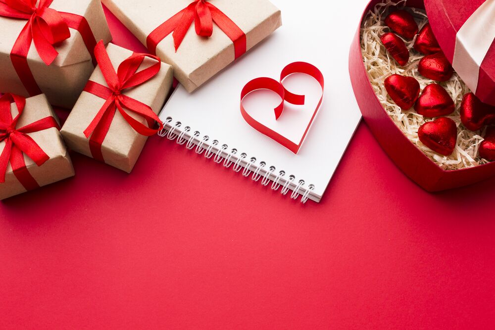 Обои для рабочего стола Конфеты в виде сердечек рядом с блокнотом и подарками на красном фоне