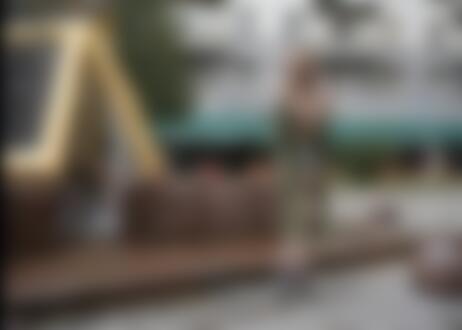 Обои на рабочий стол Светловолосая девушка в приспущенном с плеч плаще с обнаженной грудью стоит у фонтана на городской улице