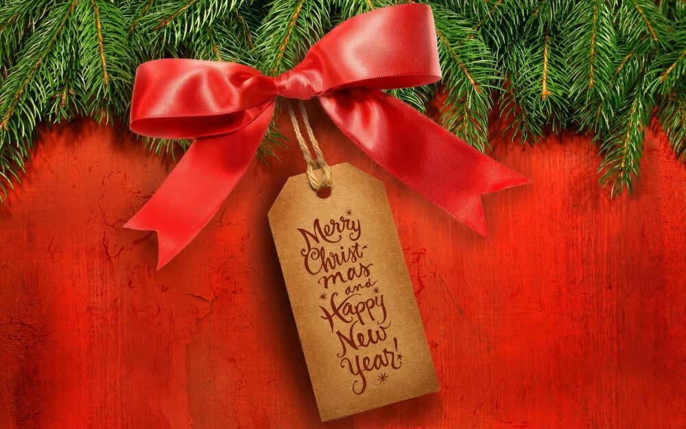 Обои для рабочего стола Ярлык с надписью «Merry Christmas and Happy New Year» с красным бантом под еловыми ветками на красном фоне