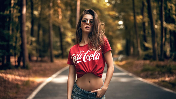 Обои на рабочий стол Молодая девушка в красной футболке с логотипом Coca-Cola и джинсовых шортах, в солнцезащитных очках, стоит на лесной дороге на фоне деревьев