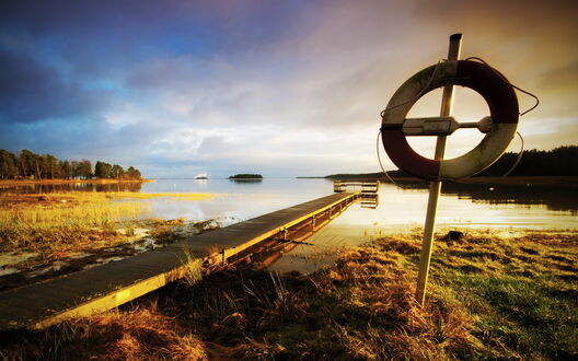 Обои на рабочий стол Деревянный пирс уходит в спокойное заросшее озеро с илистым берегом с островом вдали, на переднем плане спасательный круг на стойке, на закате