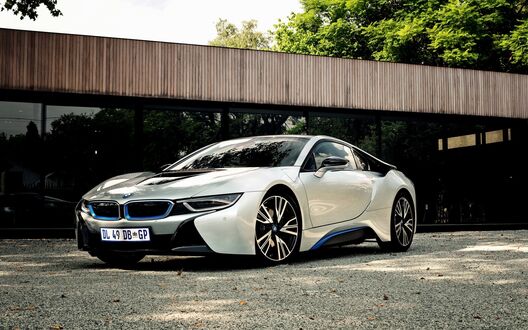 Обои на рабочий стол Белый BMW i8 с синими акцентами припаркован на гравийной площадке перед современным зданием на фоне зелени