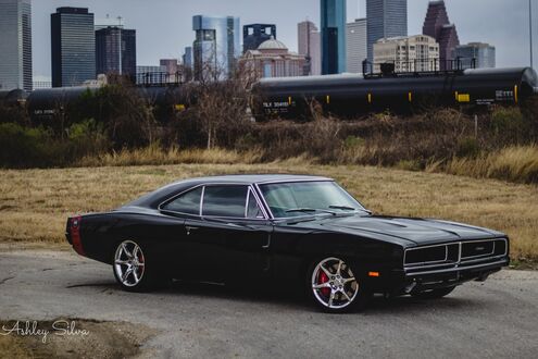 Обои на рабочий стол Черный классический muscle car Dodge Charger с хромированными дисками стоит на асфальтированной дороге на фоне грузового поезда и небоскребов Хьюстона / Houston, США, при пасмурной погоде