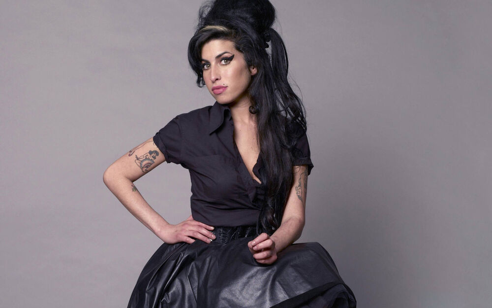 Обои для рабочего стола Эми Уайнхаус / Amy Winehouse в черной блузке и объемной юбке с высокой прической и макияжем, сидит с наклоненной головой на сером фоне