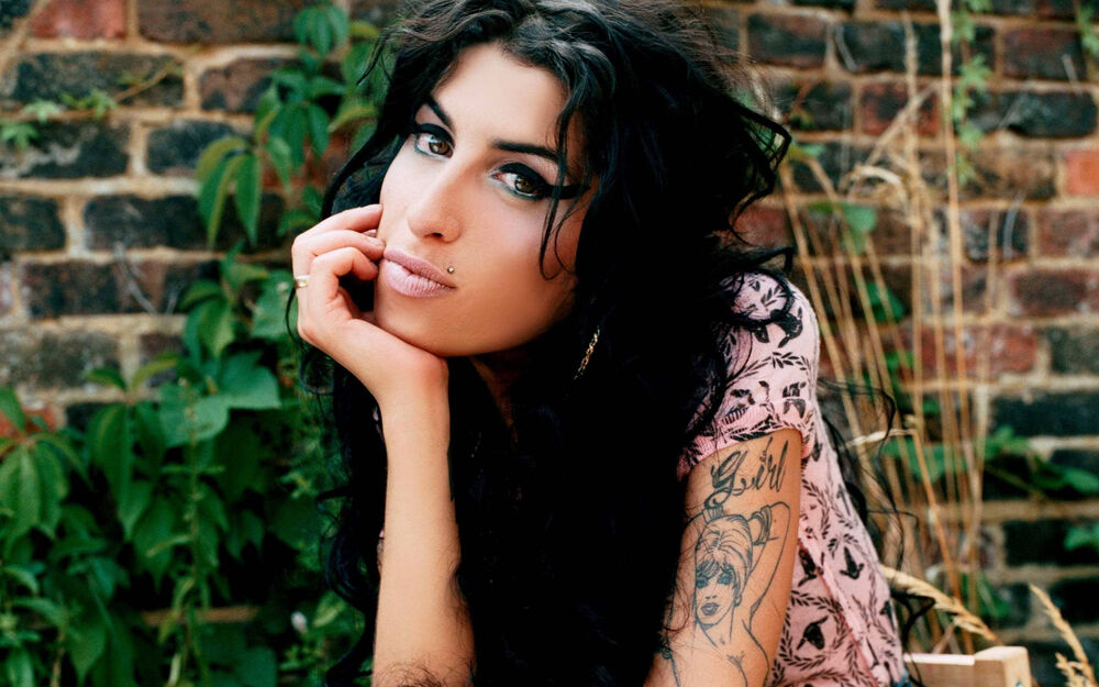 Обои для рабочего стола Эми Уайнхаус / Amy Winehouse в светлой футболке с рисунком, опершись рукой на подбородок, на фоне кирпичной стены с зелеными растениями