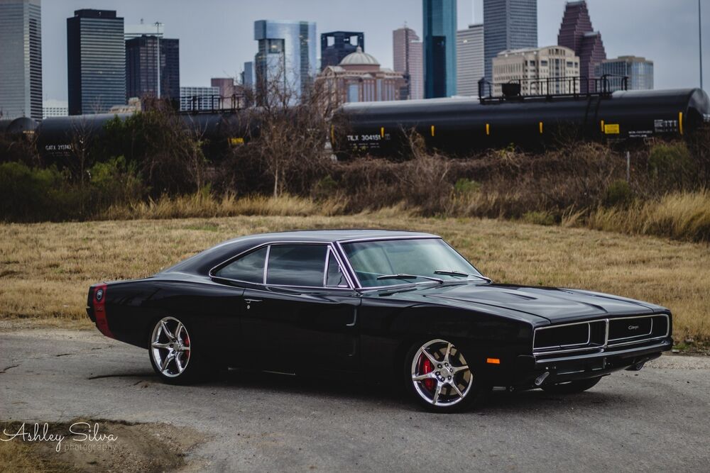 Обои для рабочего стола Черный классический muscle car Dodge Charger с хромированными дисками стоит на асфальтированной дороге на фоне грузового поезда и небоскребов Хьюстона / Houston, США, при пасмурной погоде