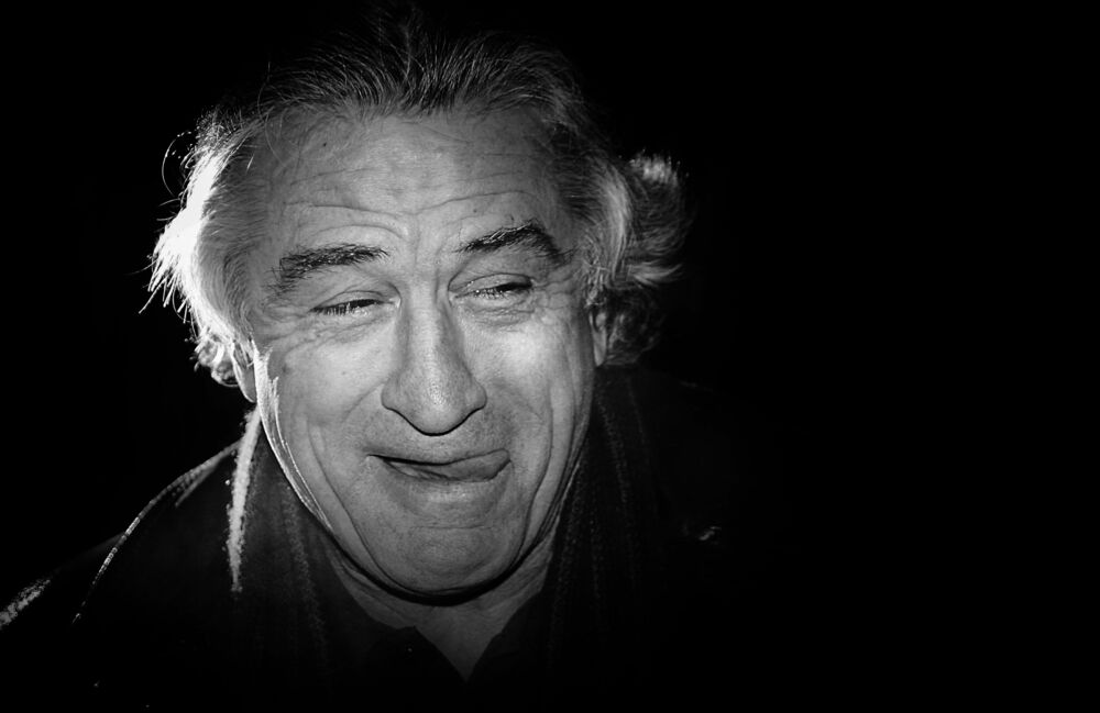 Обои для рабочего стола Роберт Де Ниро / Robert De Niro гримасничает, высунув язык на черном фоне