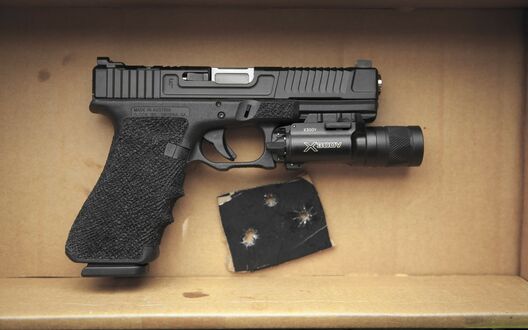 Обои на рабочий стол Черный пистолет Glock 17 с тактическим фонарем лежит в картонной коробке рядом с кусочком материала с пулевыми отверстиями