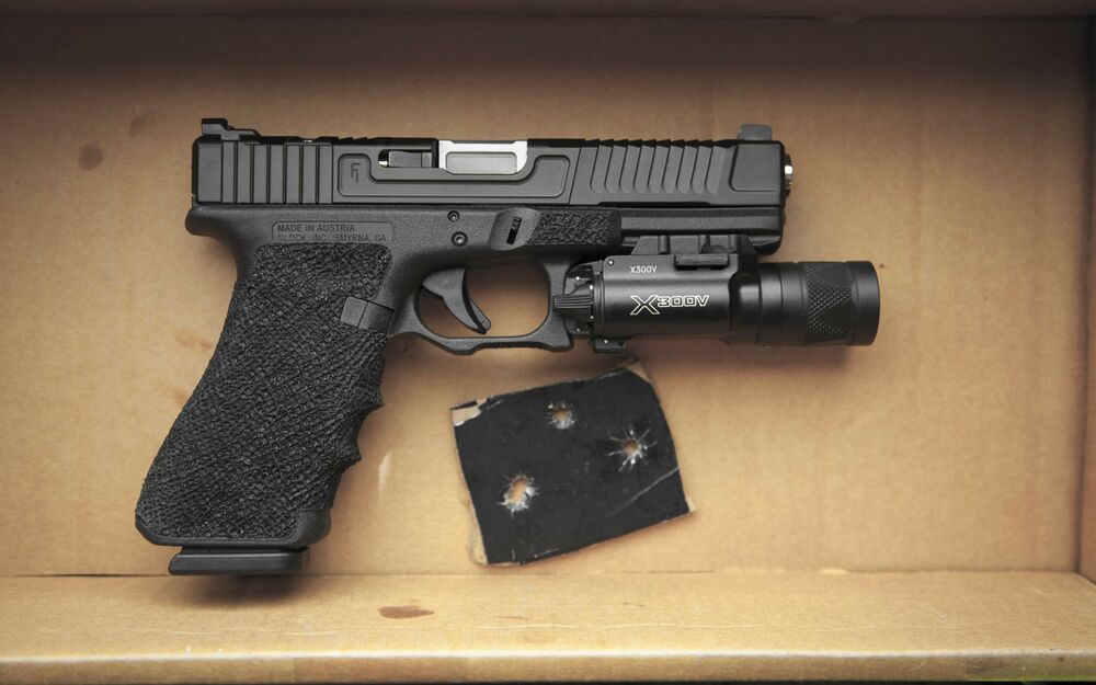 Обои для рабочего стола Черный пистолет Glock 17 с тактическим фонарем лежит в картонной коробке рядом с кусочком материала с пулевыми отверстиями