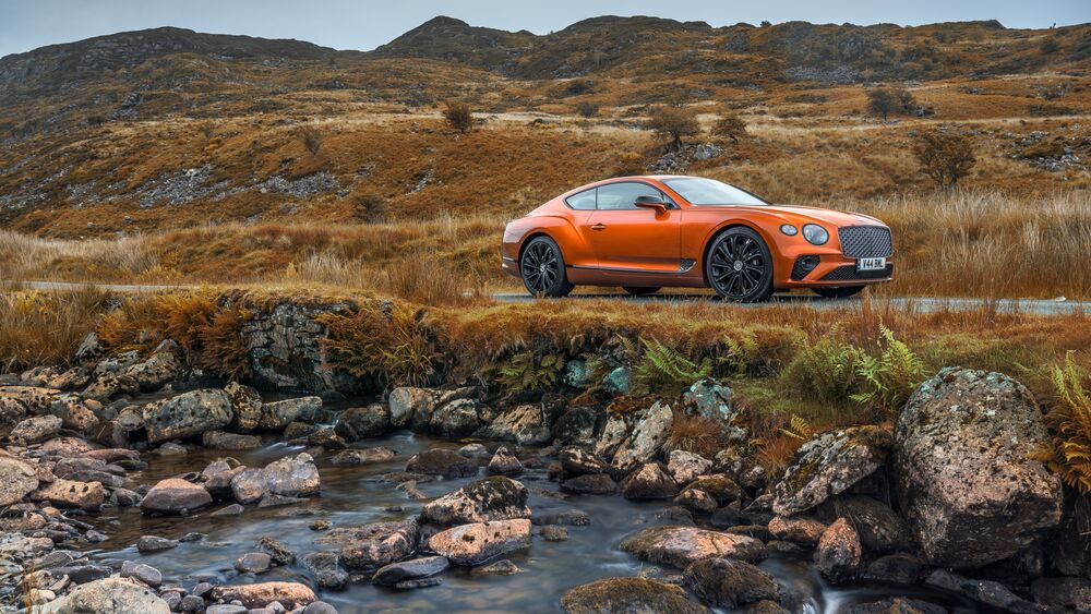 Обои для рабочего стола Bentley оранжевого цвета стоит на дороге на фоне холмов в пасмурную погоду