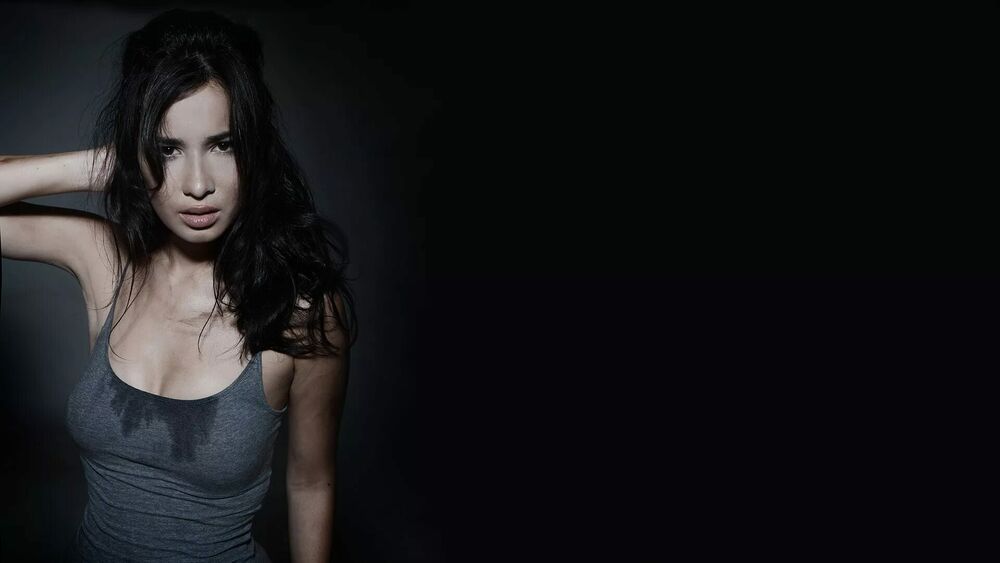 Обои для рабочего стола Селина Джейд / Celina Jade с длинными темными волосами в сером топе стоит на темном фоне