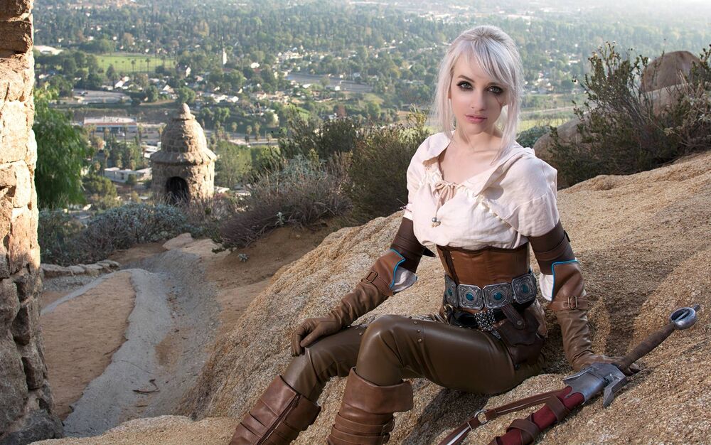 Обои для рабочего стола Цирилла / Cirilla в коричневом костюме сидит на скале на фоне горного пейзажа и каменного строения в солнечный день из игры Ведьмак 3 / The Witcher 3