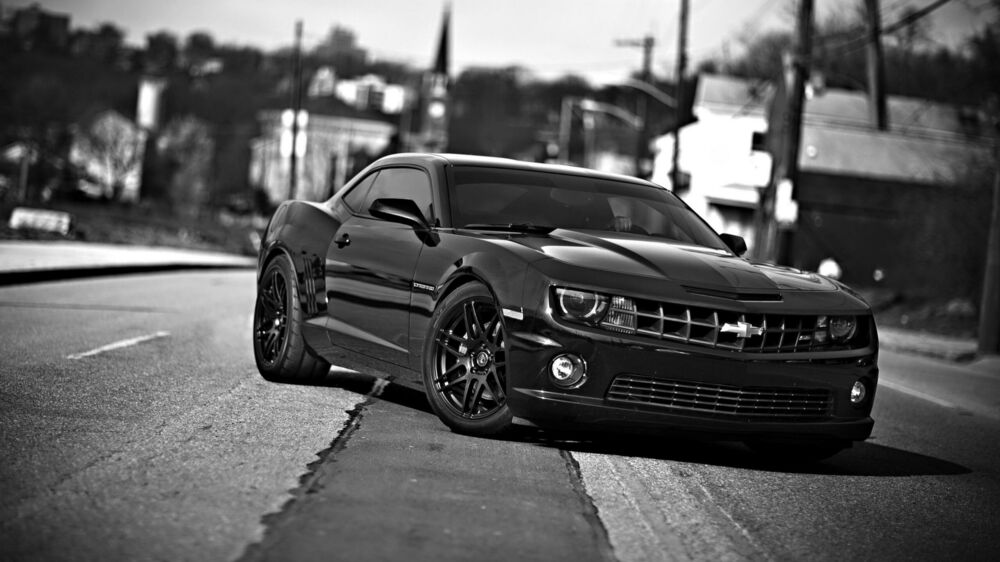 Обои для рабочего стола Chevrolet Camaro на асфальтированной дороге в городе днем на фоне размытого городского пейзажа