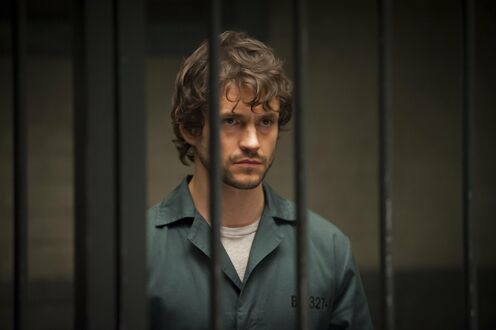 Обои на рабочий стол Мужчина с кудрявыми волосами, Уилл Грэм / Will Graham из сериала Ганнибал / Hannibal, смотрит прямо, одетый в серый тюремный костюм с надписью (B 321), на фоне темной стены и тюремных решеток