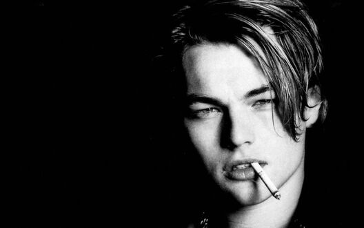 Обои на рабочий стол Молодой Леонардо Ди Каприо / Leonardo DiCaprio, с влажной уложенной челкой держит сигарету между губ и смотрит прямо, на черном фоне