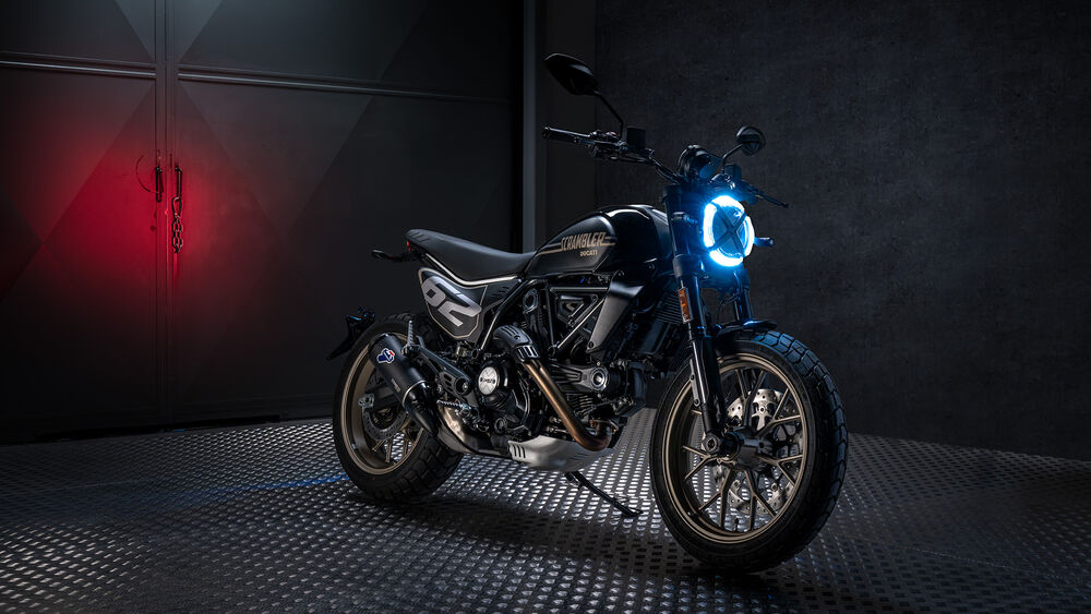 Обои для рабочего стола Мотоцикл Ducati Scrambler черного цвета с ярко-синей круглой фарой стоит на металлической поверхности на темном фоне с красным свечением