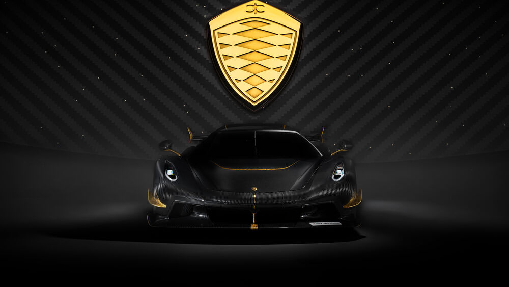 Обои для рабочего стола Черный Koenigsegg Jesko c золотыми акцентами стоит, на фоне темной стены с диагональными полосами и золотым логотипом Koenigsegg
