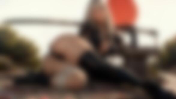 Обои на рабочий стол Девушка в черном костюме и высоких сапогах позирует на каменных плитах, держа катану, на фоне красного круга в небе и зеленых деревьев с розовыми лепестками