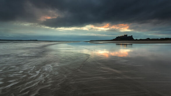 Обои на рабочий стол Замок Бамбург / Bamburgh Castle, Великобритания / United Kingdom, мокрый песчаный пляж с отражениями неба, на фоне силуэта замка и облачного неба с оранжевыми и серыми оттенками