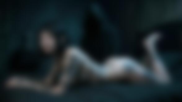 Обои на рабочий стол Молодая женщина с татуировками на теле позирует, лежа на животе в черном белье, на фоне темной комнаты с фигурой в черном капюшоне и крестом